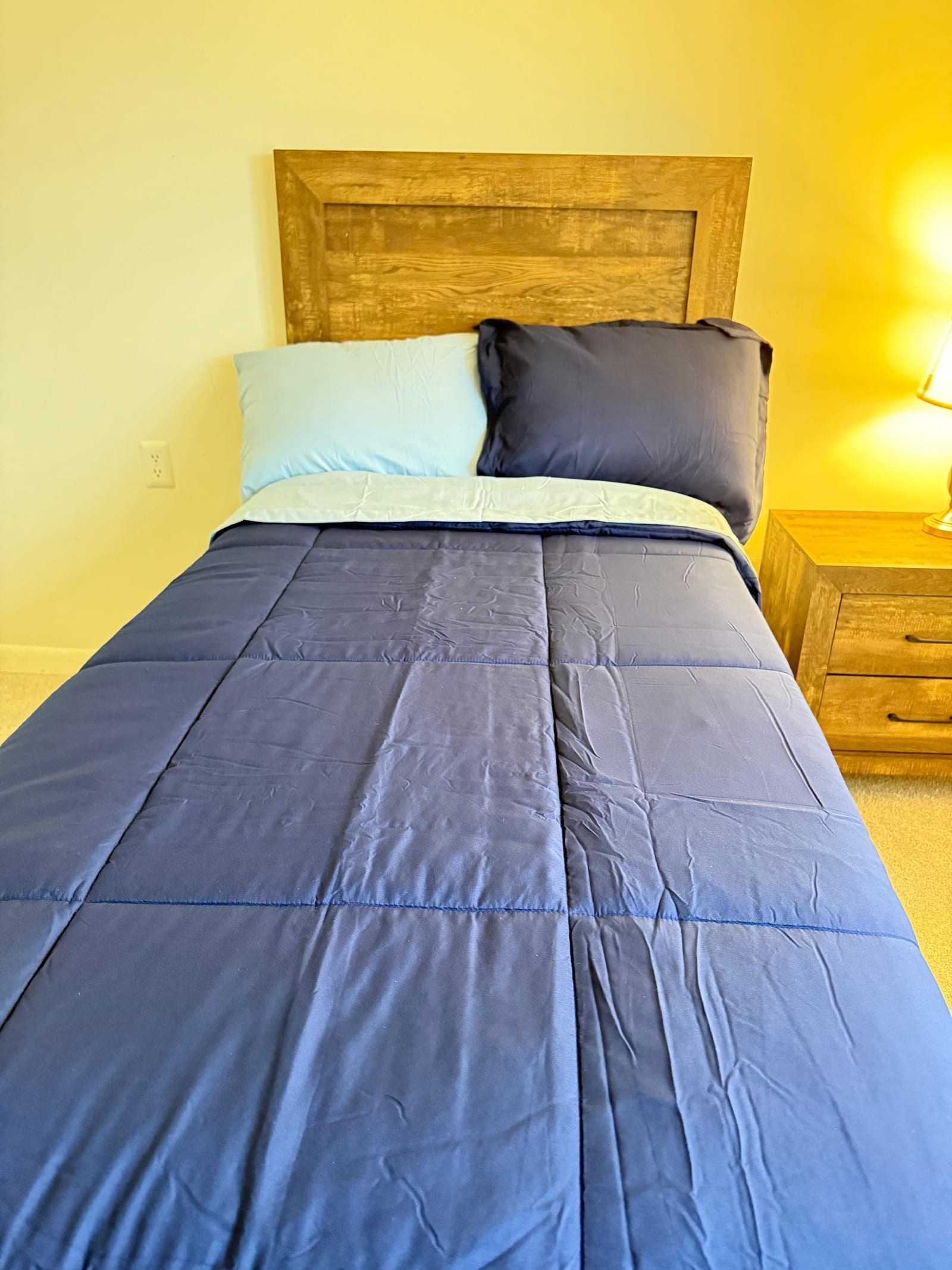 A private bedroom with a made single bed, a wooden headboard and a bedside table with a lamp.
