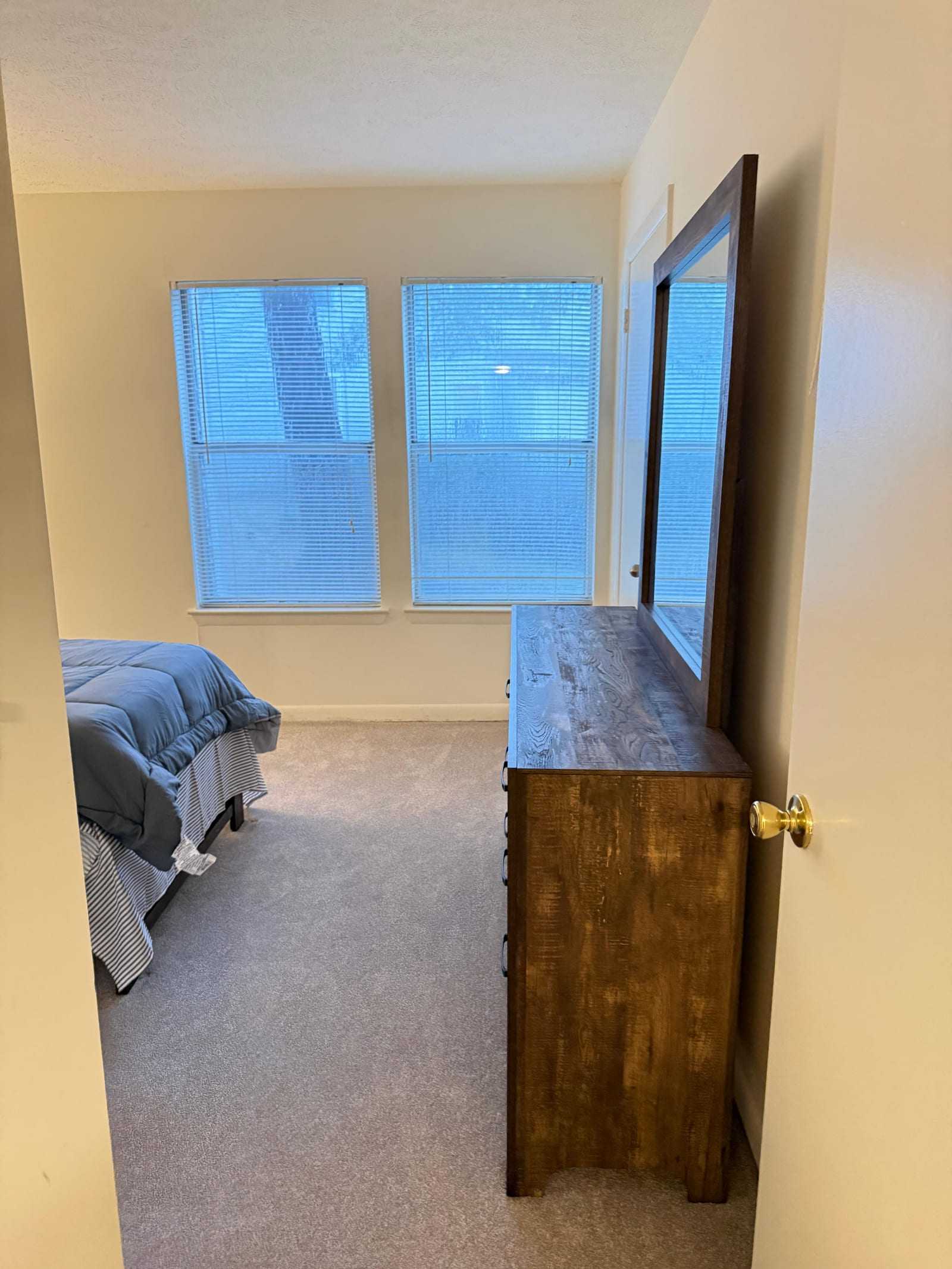 A private bedroom with two curtained windows, a made bed and a tall wooden dresser with a mirror.