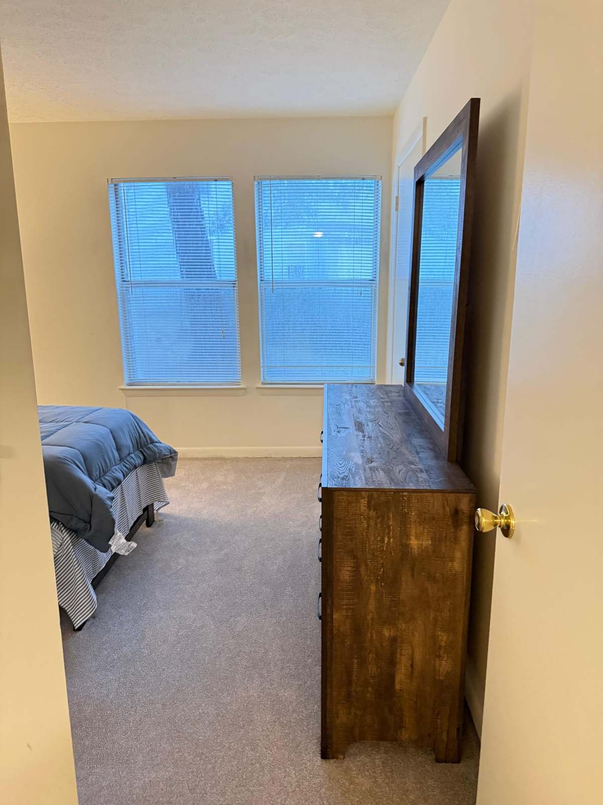 A private bedroom with two curtained windows, a made bed and a tall wooden dresser with a mirror.