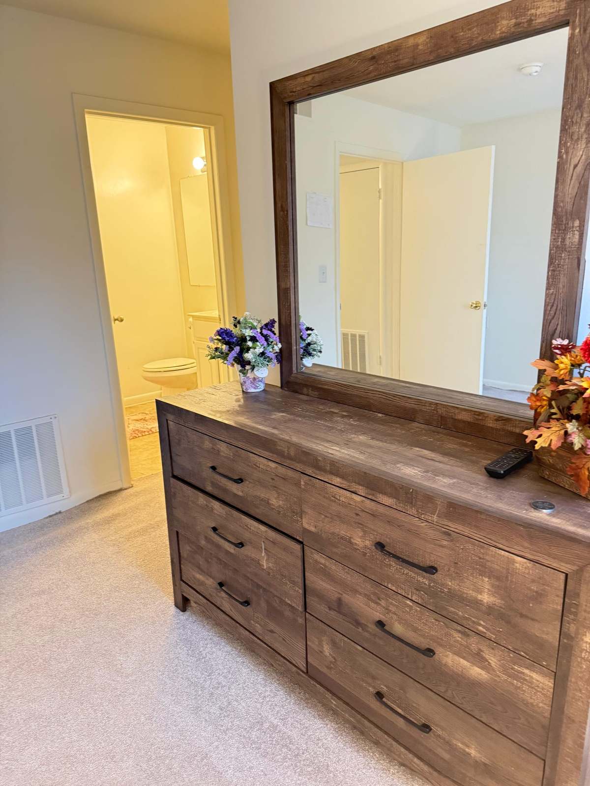 A bedroom dresser with a large mirror and fresh flowers, with the door to the en-suite bathroom open beyond.