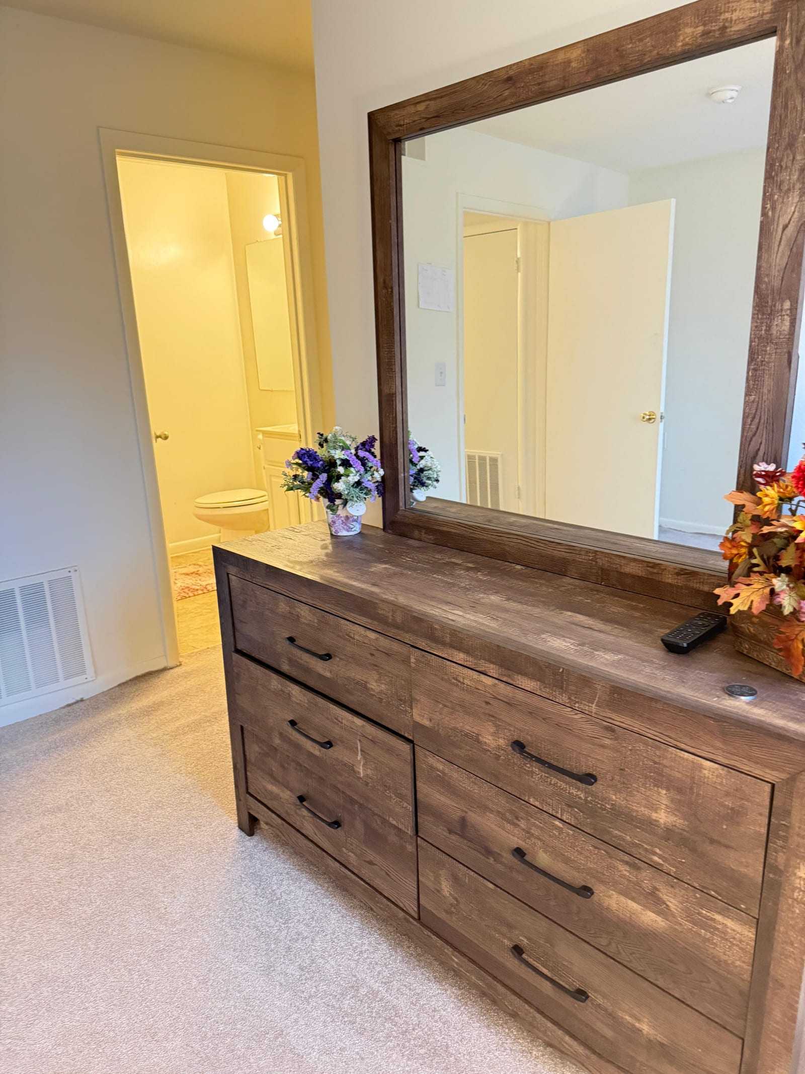 A bedroom dresser with a large mirror and fresh flowers, with the door to the en-suite bathroom open beyond.