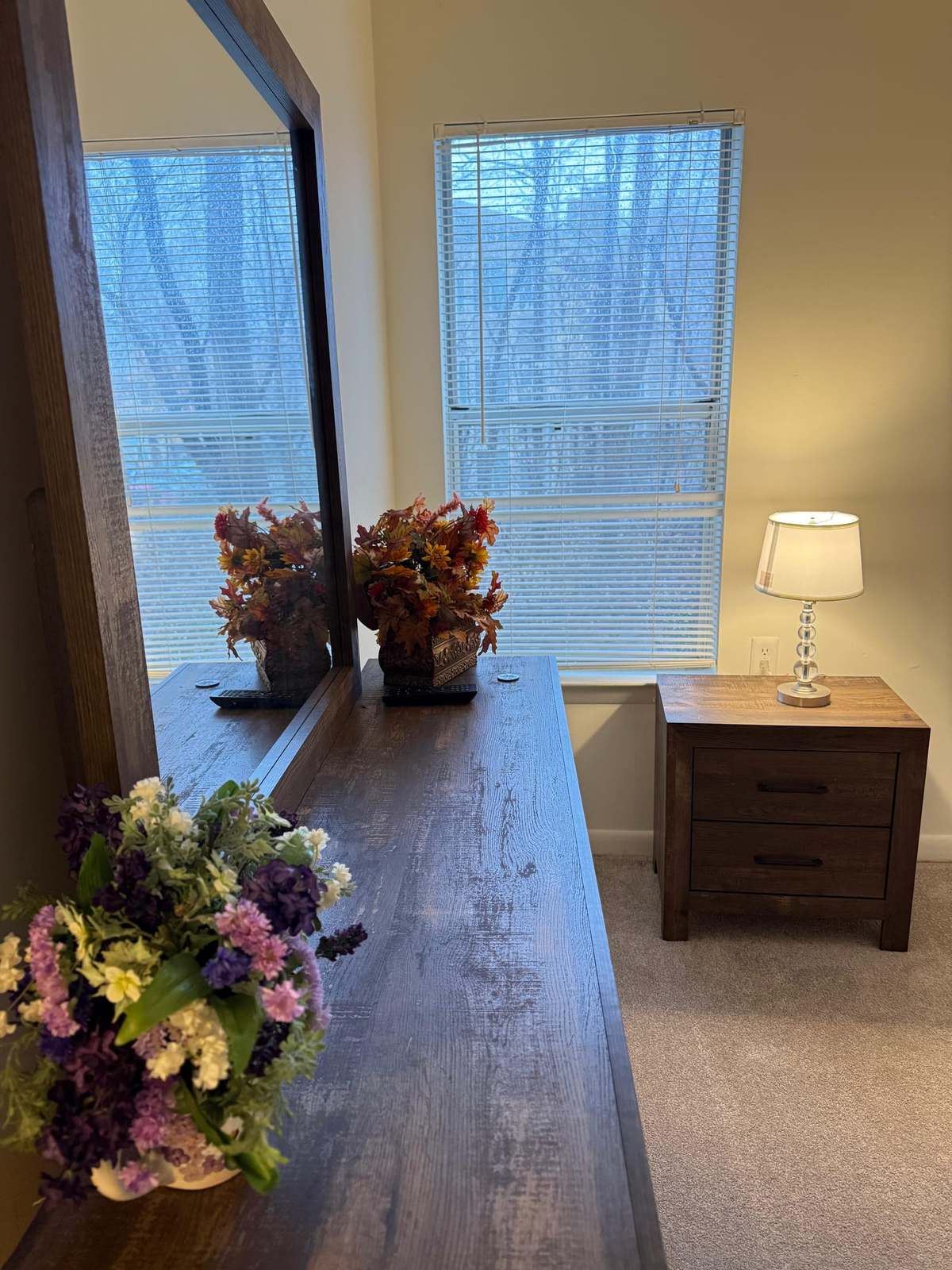 A bedroom dresser with a mirror and flowers beside a window, with a bedside table and lamp against the far wall.