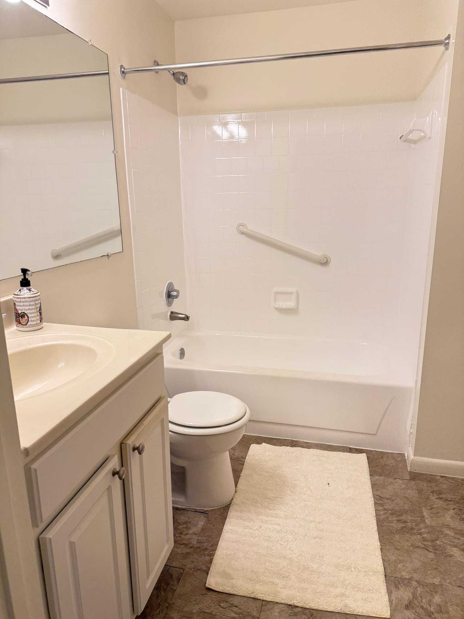 A full bathroom with a grab bar fitted along the wall of the bath, a vanity unit with mirror, and a non-slip mat.