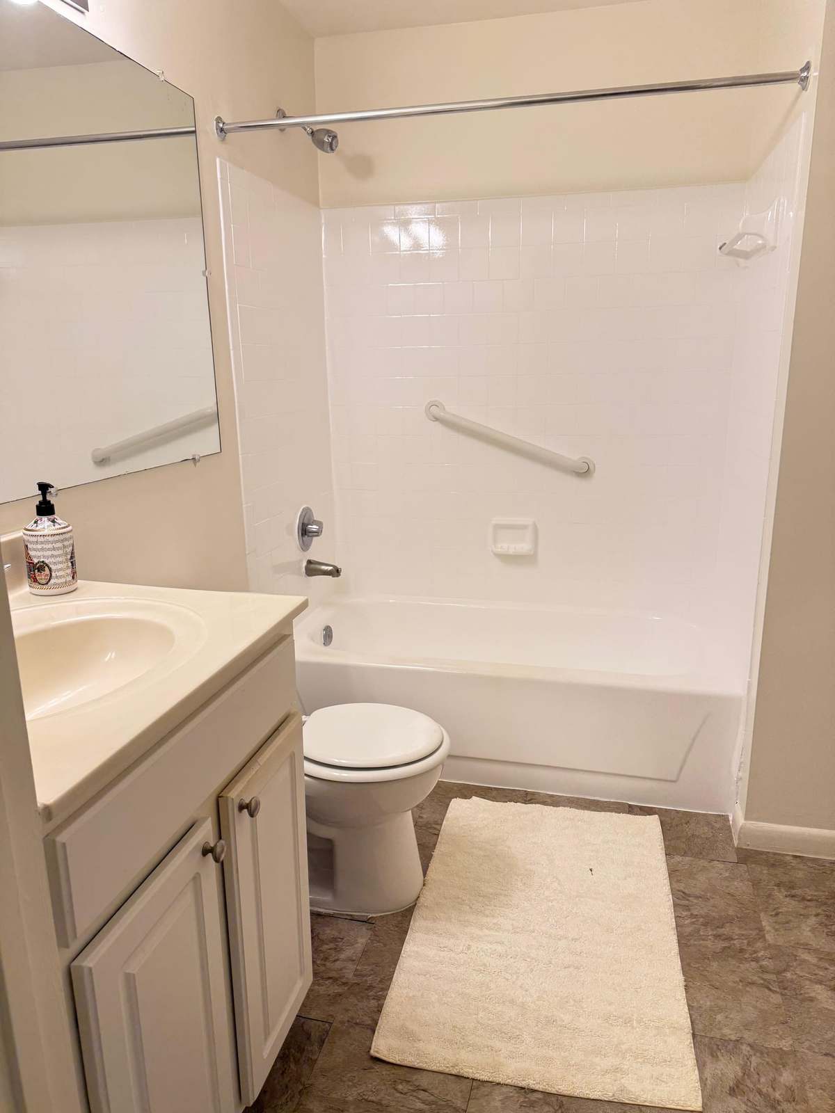 A full bathroom with a grab bar fitted along the wall of the bath, a vanity unit with mirror, and a non-slip mat.