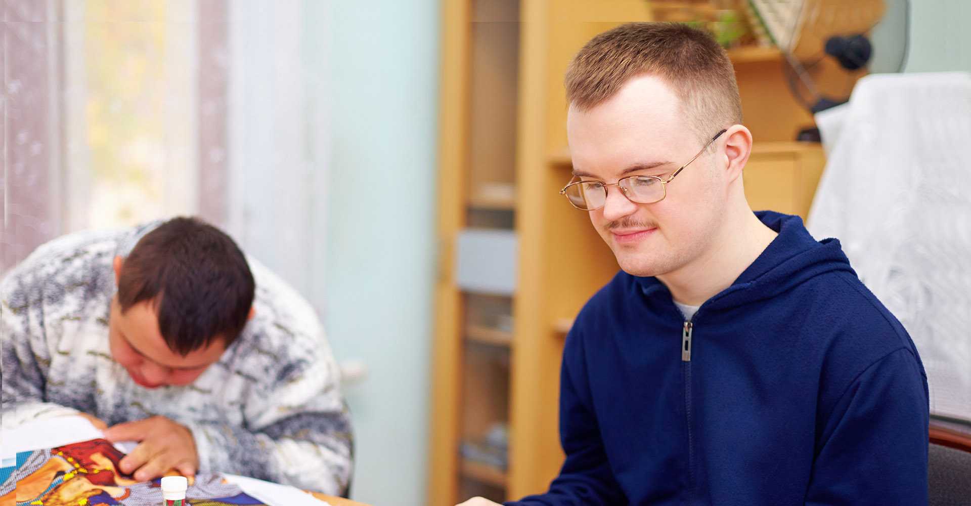 Two men with developmental disabilities working on a craft project together at a table in a bright living room.
