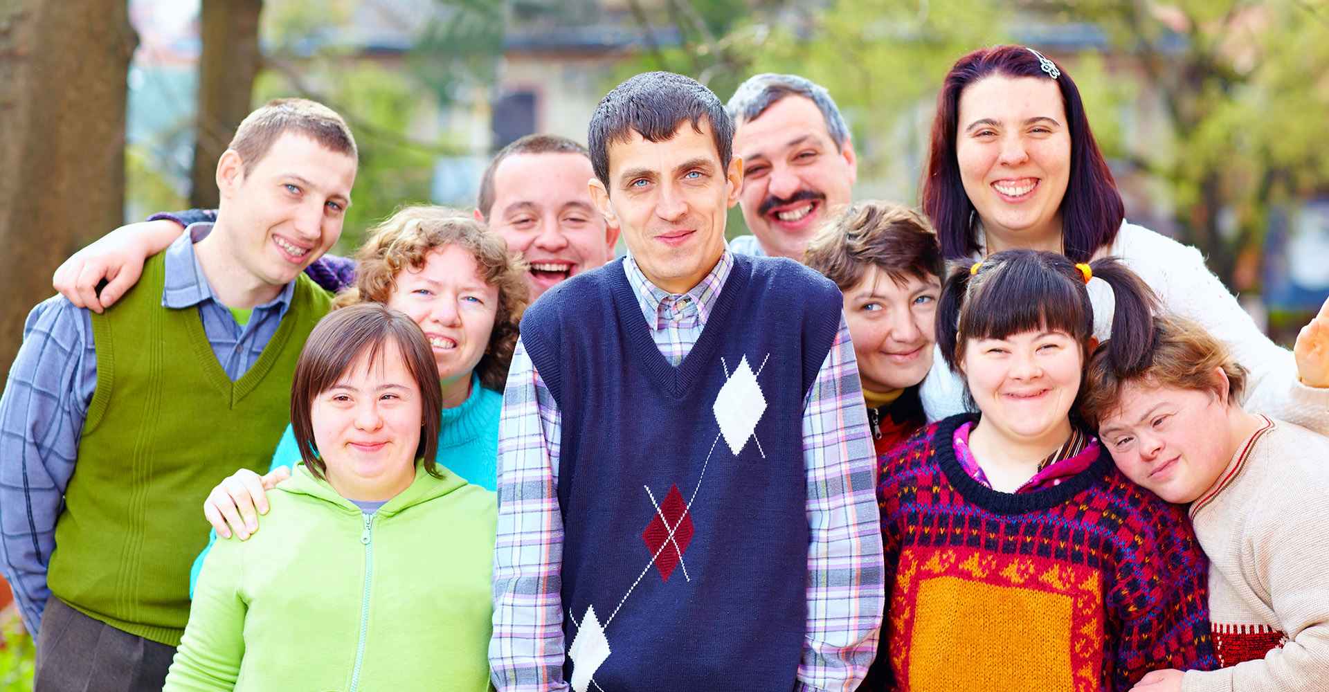 A group of adults with developmental disabilities standing together outdoors, smiling with arms around each other.