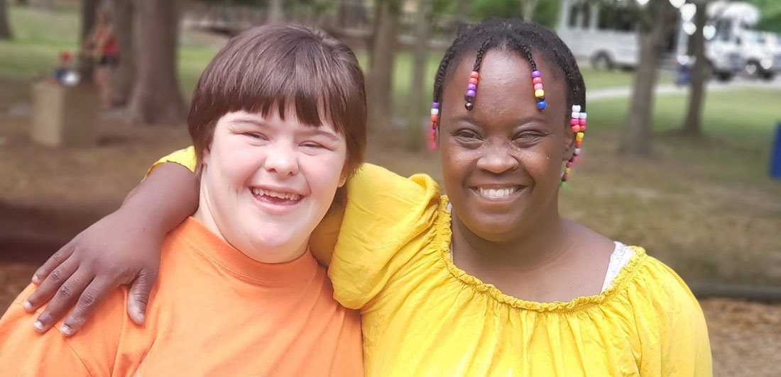Two young women, one with Down syndrome, standing arm in arm outdoors and smiling at the camera.