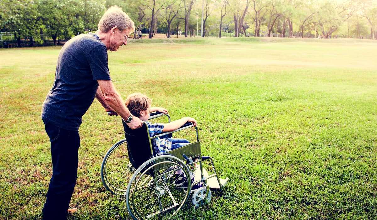 A support worker walking alongside a young person using a wheelchair on a path through a green park.